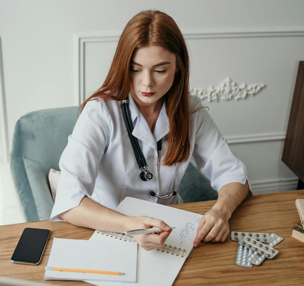 pexels-photo-5407206-5407206 A female doctor writes notes at her desk in a clinical setting, focusing on healthcare tasks.
